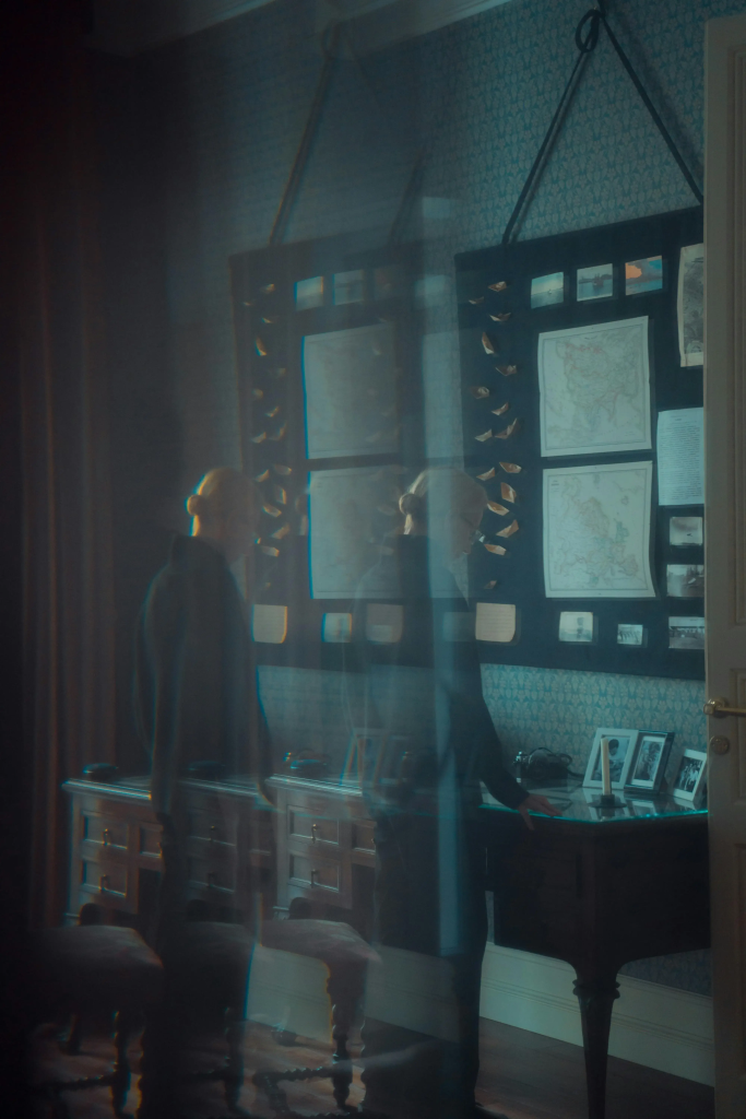 A woman reflected in glass as she stands in a nostalgic room filled with old maps, photographs, and memorabilia.