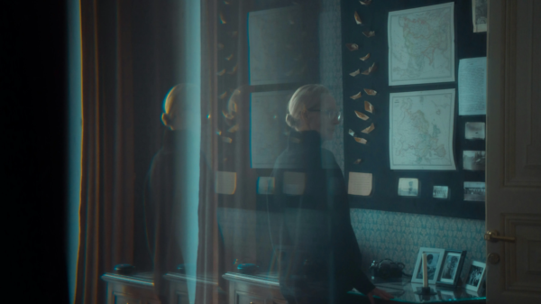 A vintage study room with maps, photos, and collectible items on the wall, captured with a blurred silhouette of a person passing by.