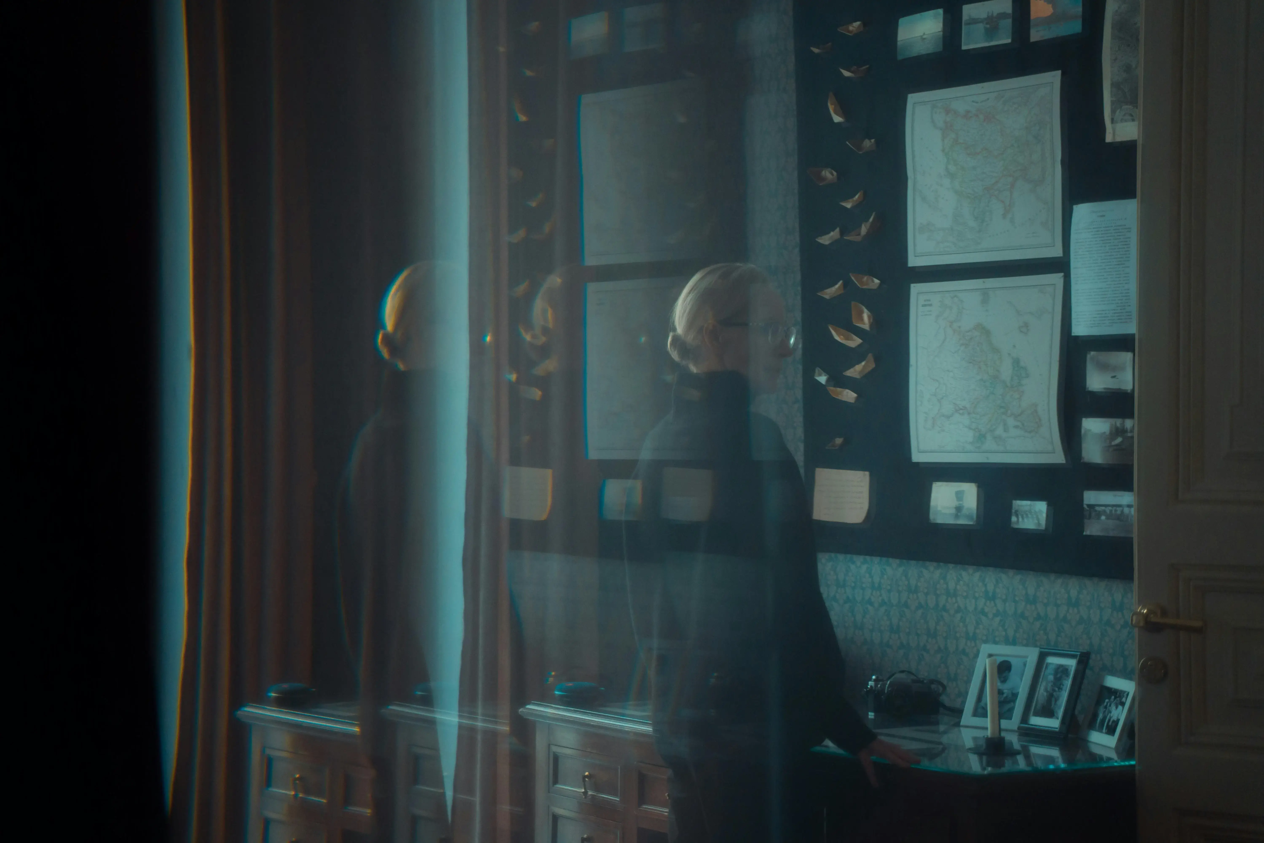 A vintage study room with maps, photos, and collectible items on the wall, captured with a blurred silhouette of a person passing by.