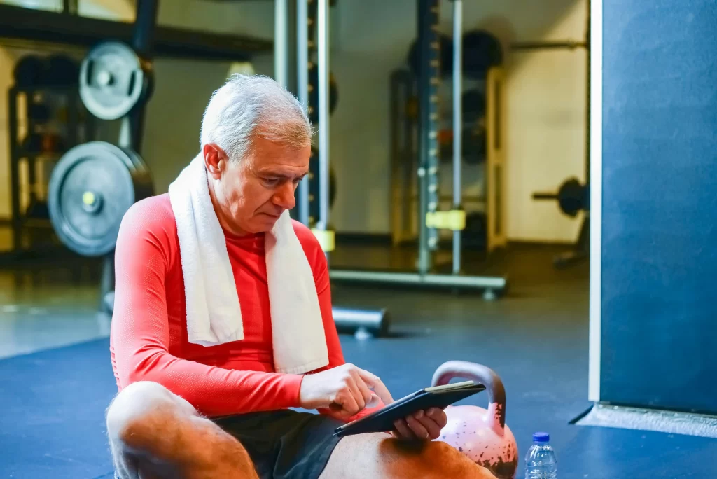 Older man sitting on the gym floor checking his tablet after a workout, showing how to train your body for your second act.