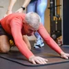 Older man stretching on a gym mat with a trainer’s support, demonstrating mobility training for your second act.