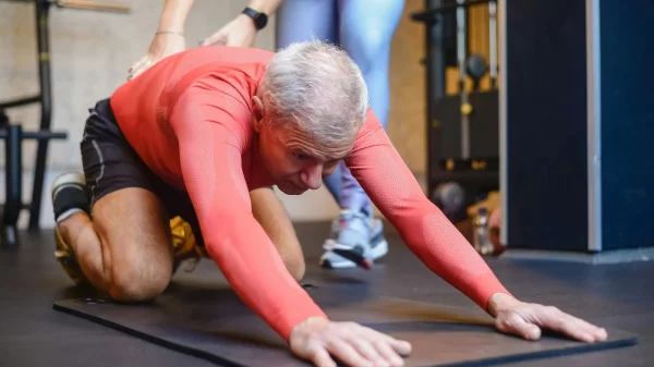 Older man stretching on a gym mat with a trainer’s support, demonstrating mobility training for your second act.