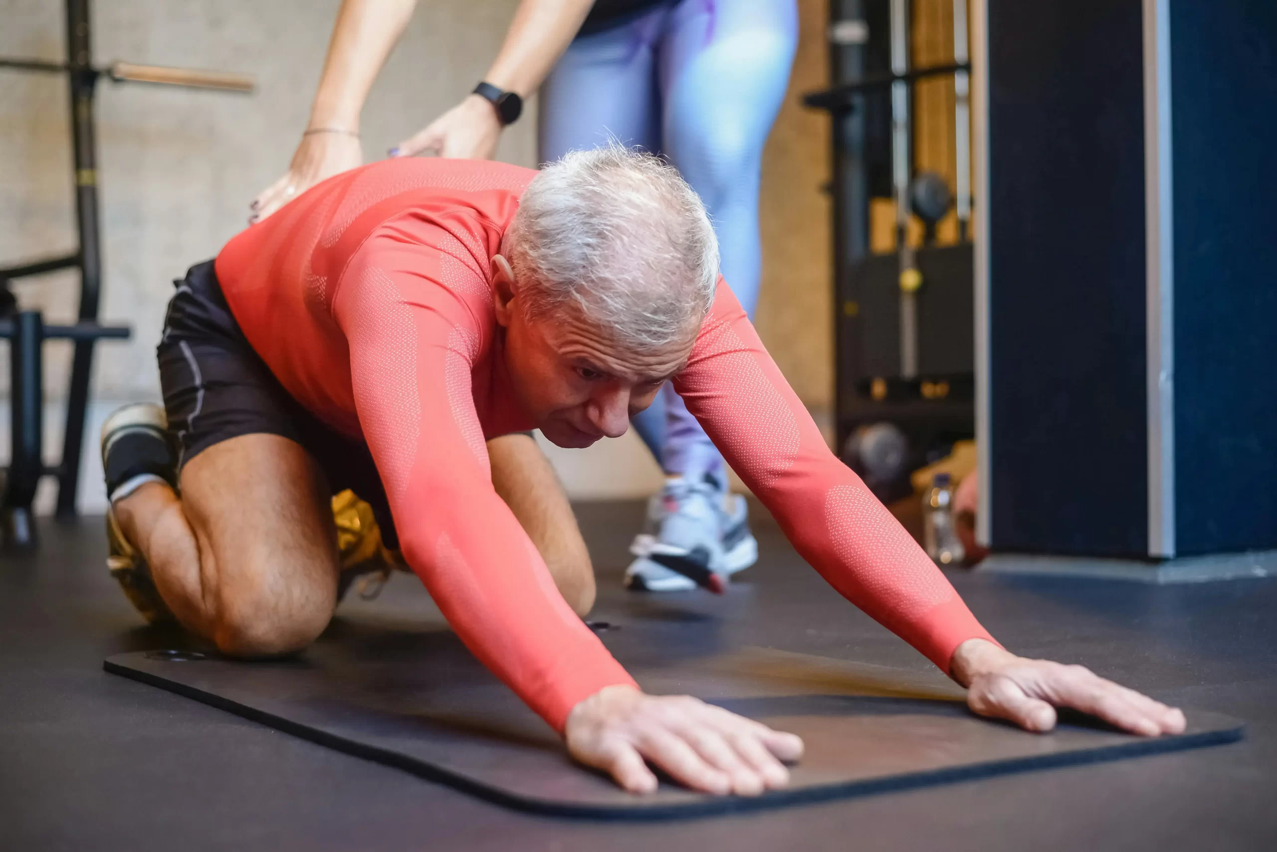 Older man stretching on a gym mat with a trainer’s support, demonstrating mobility training for your second act.