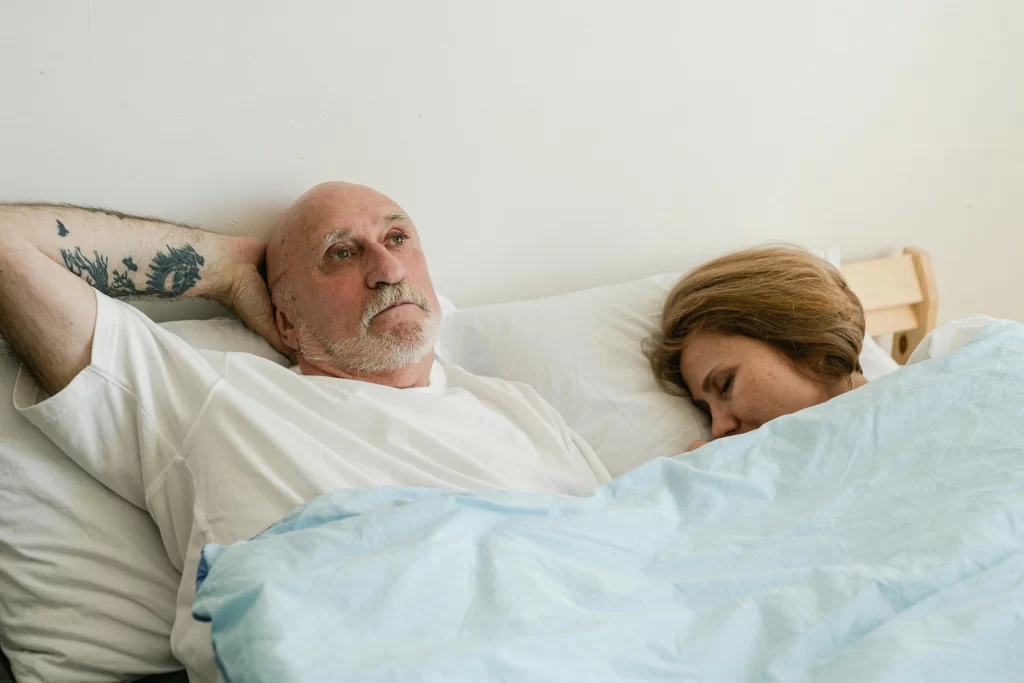 Older man awake in bed beside his sleeping partner, showing how sleep hits different after 45.