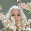 Older woman with long white hair smiling gently while surrounded by soft pink flowers.