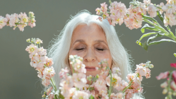 Older woman with long white hair smiling gently while surrounded by soft pink flowers.