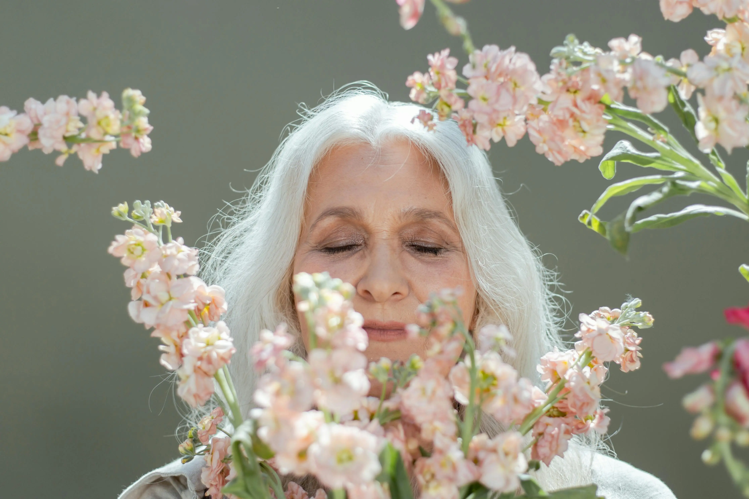 Older woman with long white hair smiling gently while surrounded by soft pink flowers.