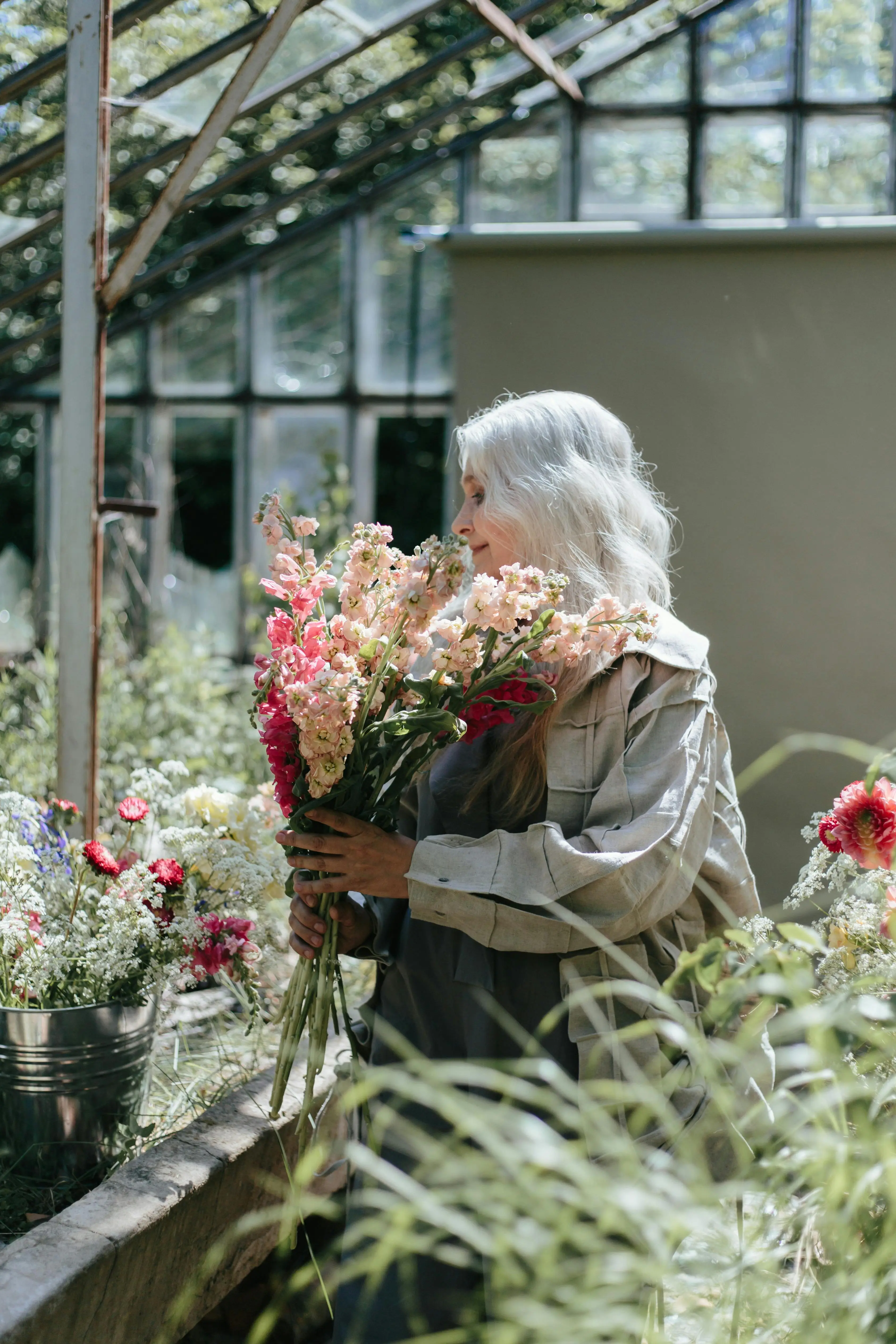 Mature woman with white hair holding a bouquet of colorful flowers inside a greenhouse.