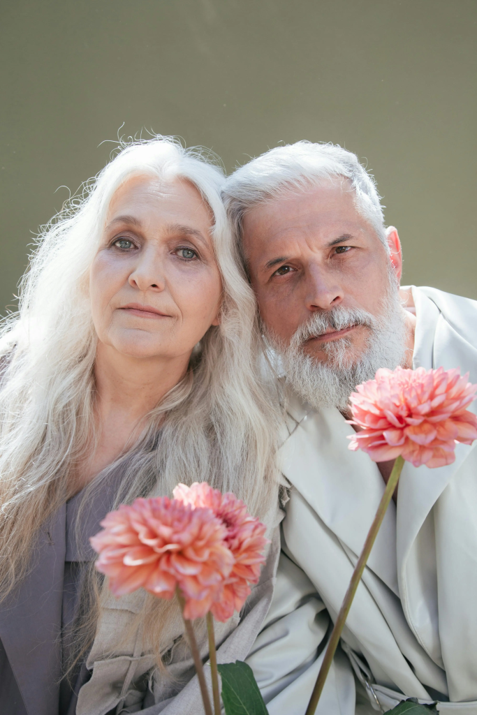 Older couple with white hair posing closely together and holding pink flowers.