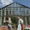 Older couple standing in front of a vintage greenhouse, talking while holding flowers