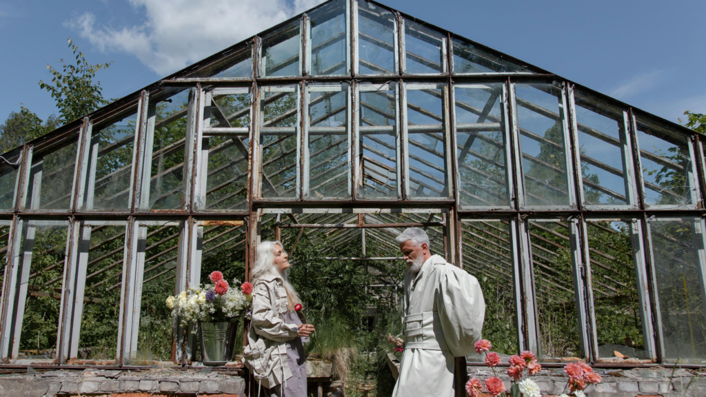 Older couple standing in front of a vintage greenhouse, talking while holding flowers