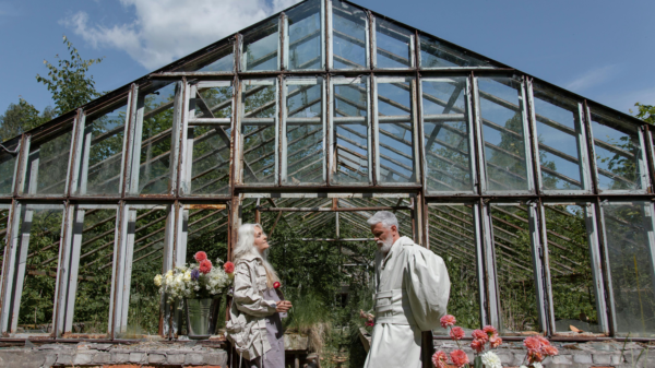 Older couple standing in front of a vintage greenhouse, talking while holding flowers
