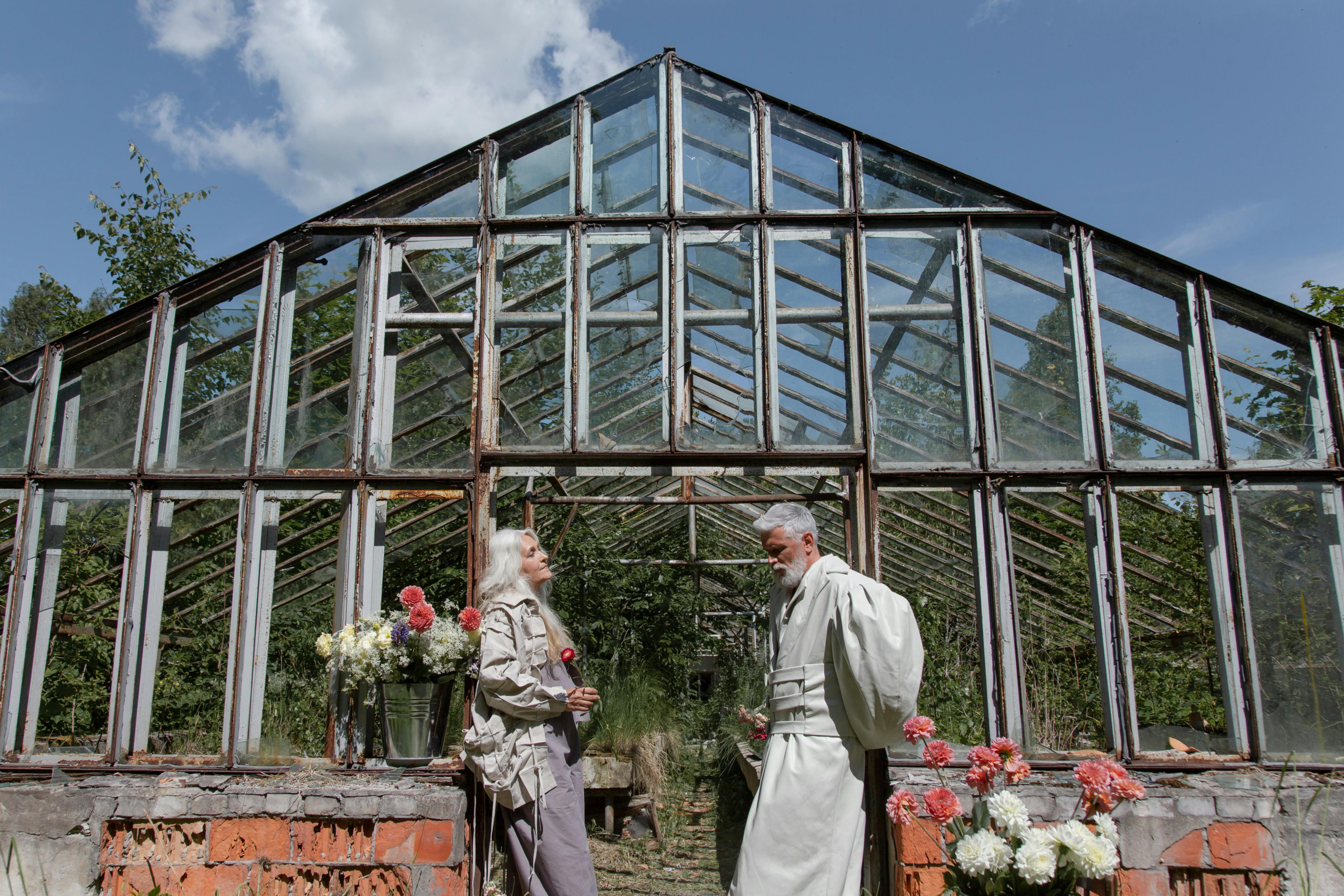 Older couple standing in front of a vintage greenhouse, talking while holding flowers