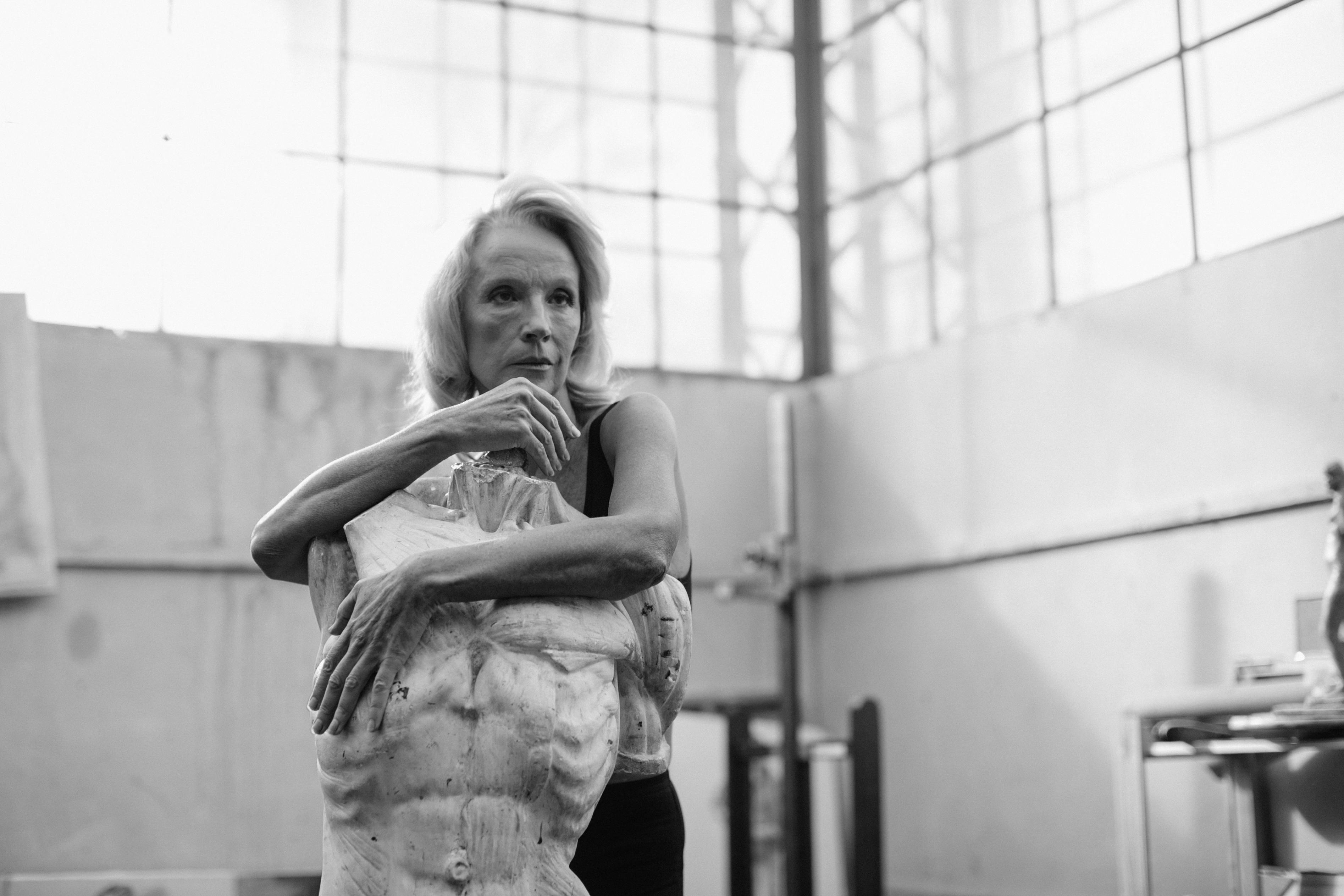 Mature woman in an art studio leaning on a sculpted torso, appearing thoughtful and focused.