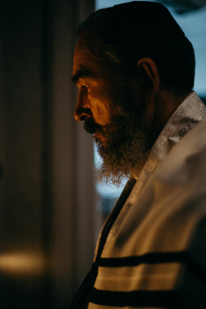 Side profile of an older man with a beard standing in warm candlelight, reflecting quietly.