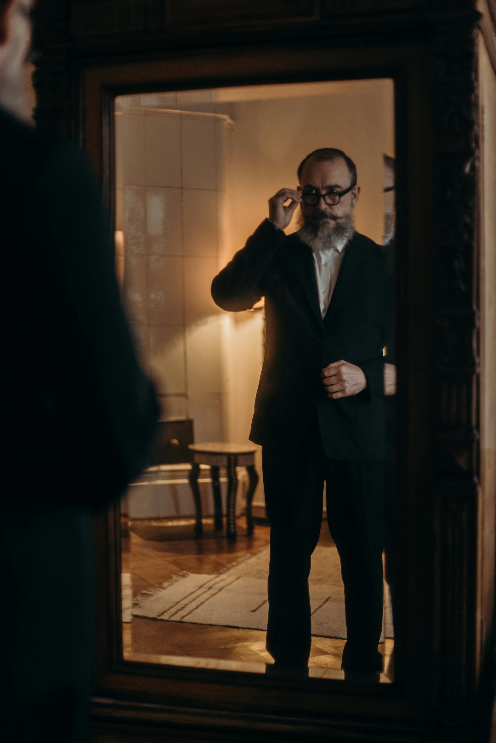 Man in his 40s looking at his reflection while adjusting glasses, symbolizing body awareness and aging