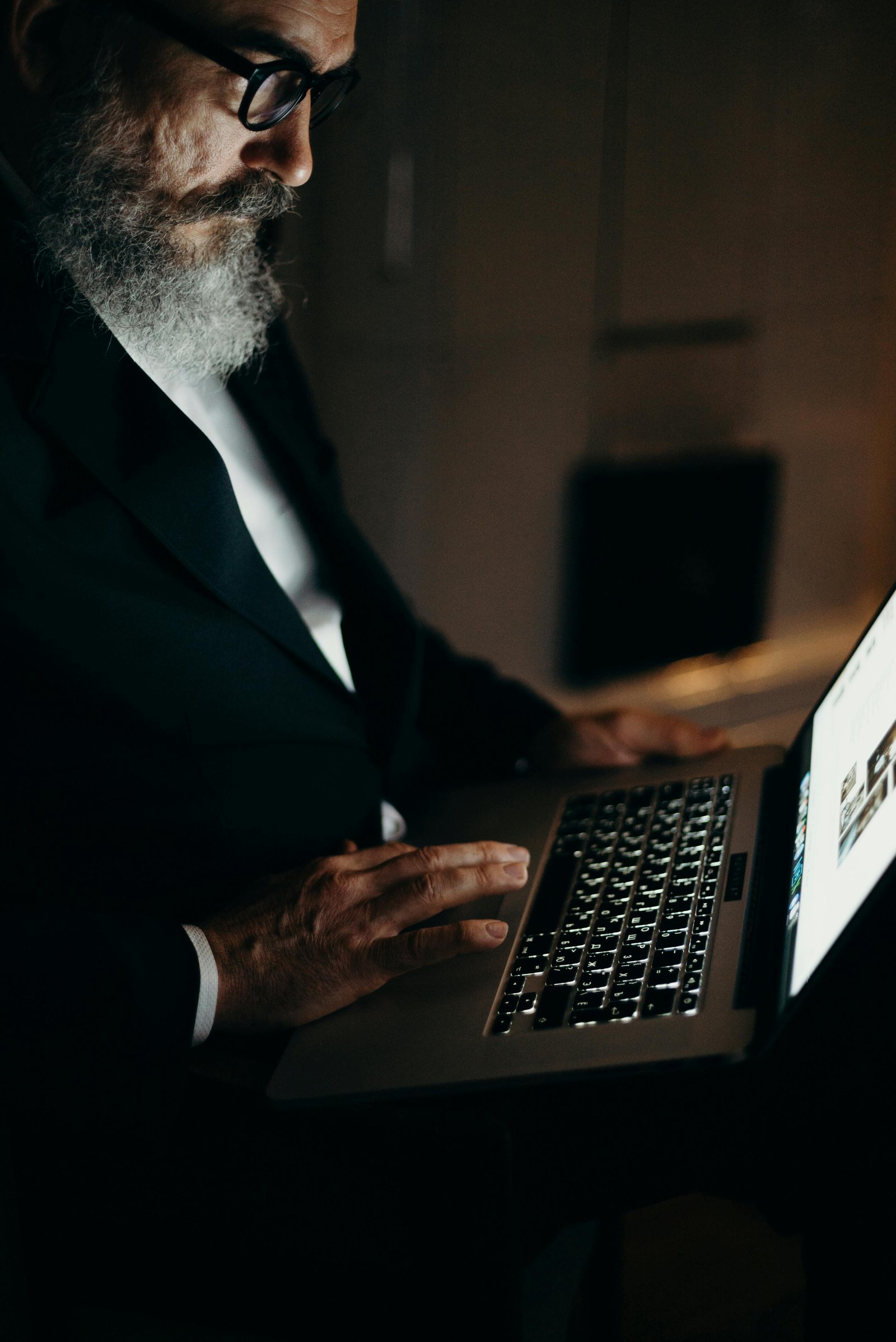 Close-up of an older man with a gray beard and glasses focusing on his laptop as he types in a dark room.