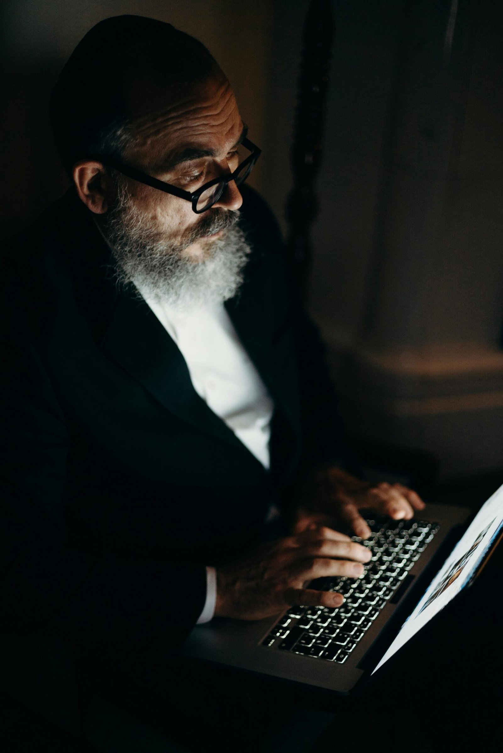 Bearded older man wearing glasses and a suit, typing on a laptop illuminated by its screen in a dark room.