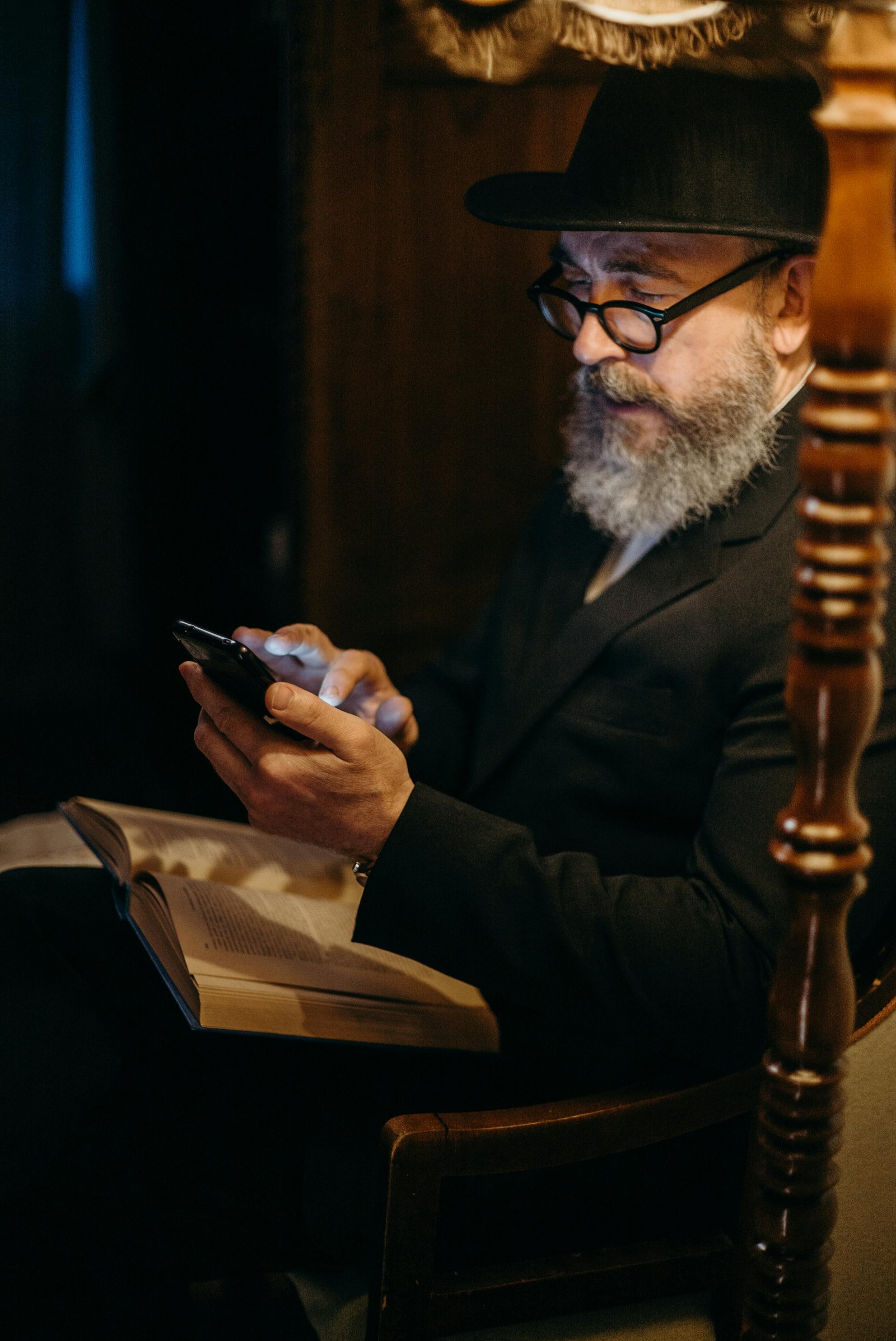 Older man with a long gray beard and glasses sitting in a dimly lit room, using a smartphone while an open book rests on his lap.