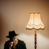 Older man in a black suit and hat sitting beside a vintage lamp, deep in thought in a dimly lit room.
