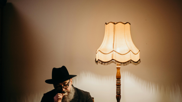 Older man in a black suit and hat sitting beside a vintage lamp, deep in thought in a dimly lit room.