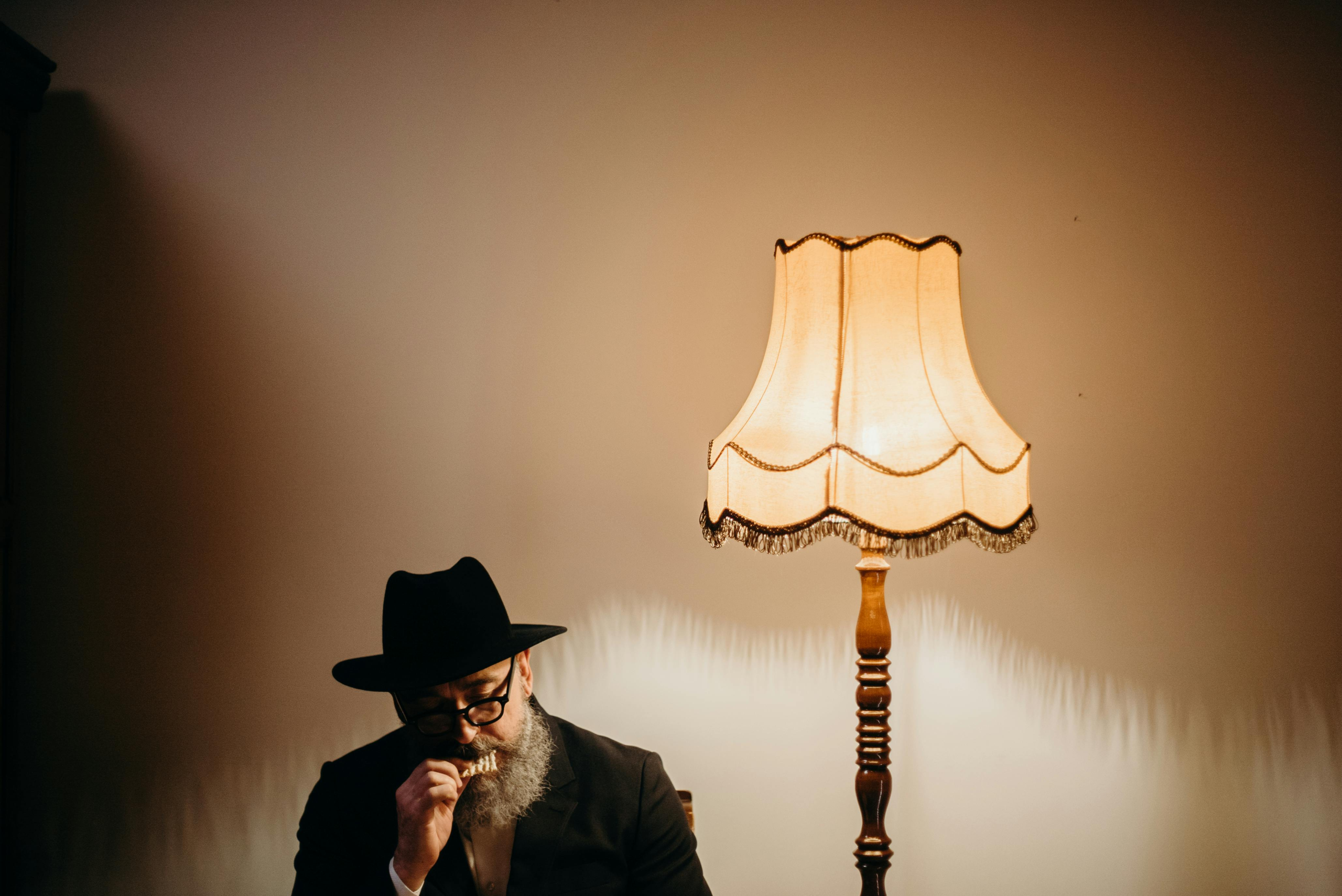 Older man in a black suit and hat sitting beside a vintage lamp, deep in thought in a dimly lit room.