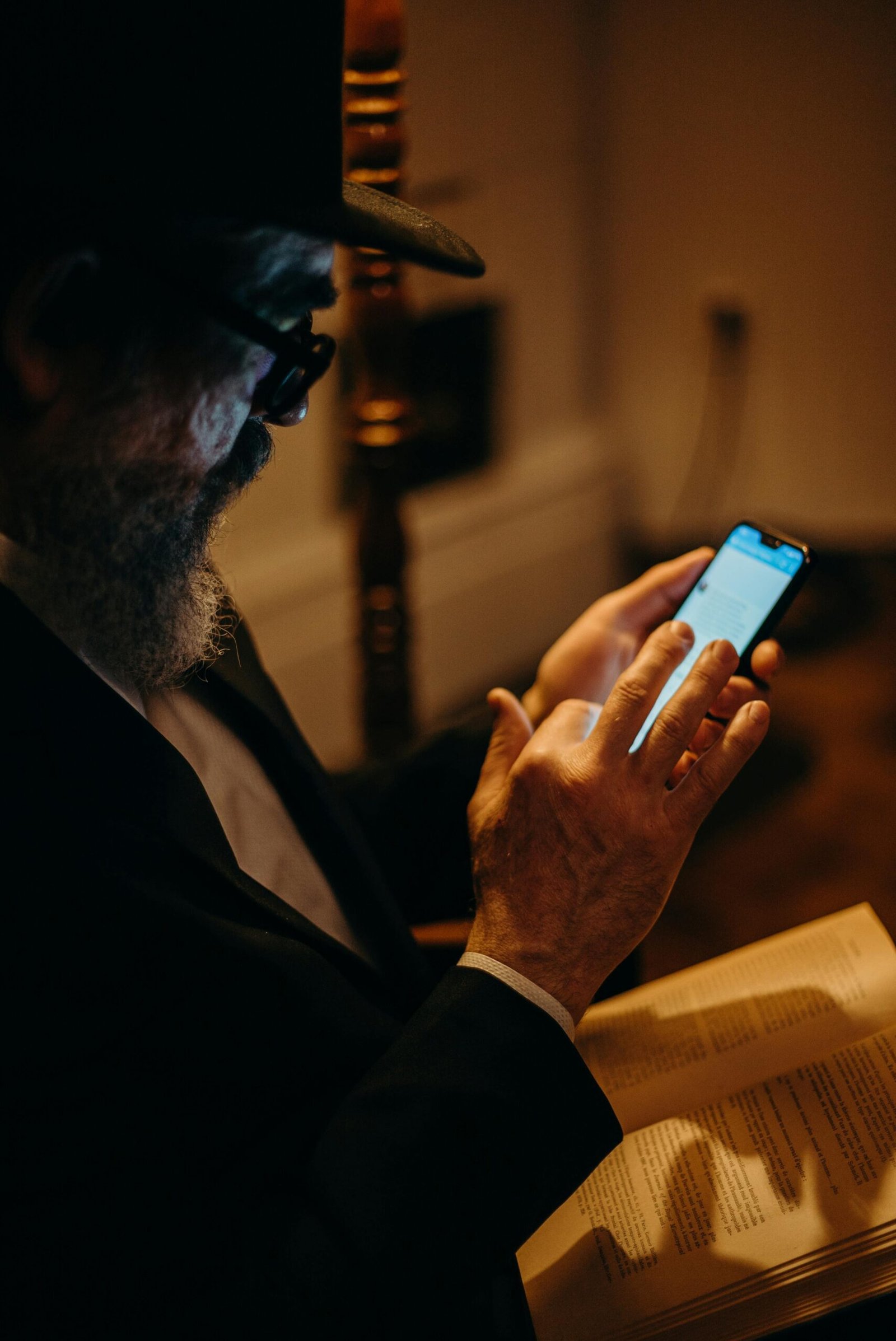 Older man wearing glasses and a hat using a smartphone while holding an open book.