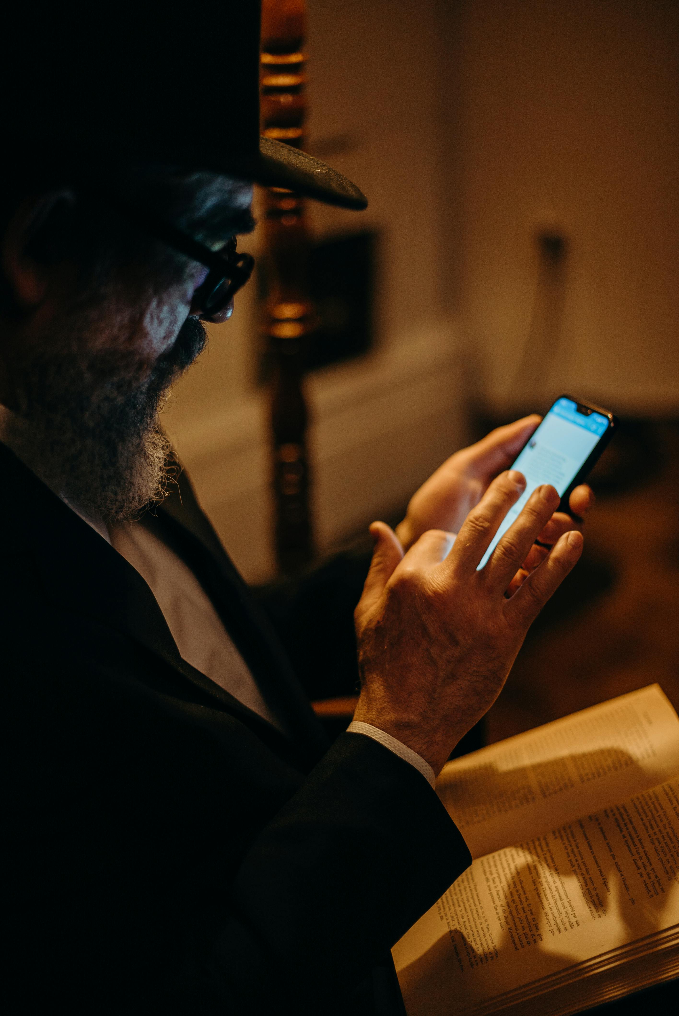 Older man wearing glasses and a hat using a smartphone while holding an open book.