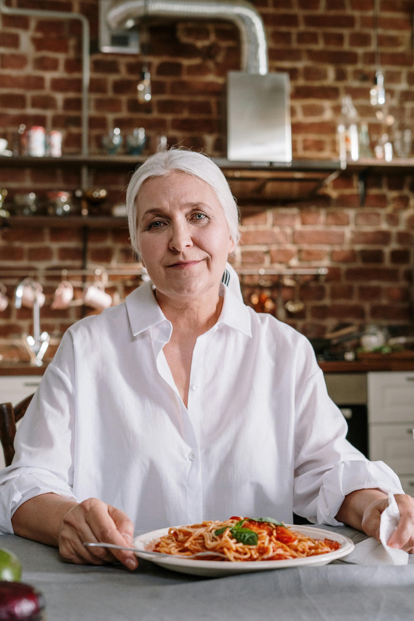 Older woman with white hair sitting in a cozy kitchen, smiling gently while enjoying a plate of pasta.