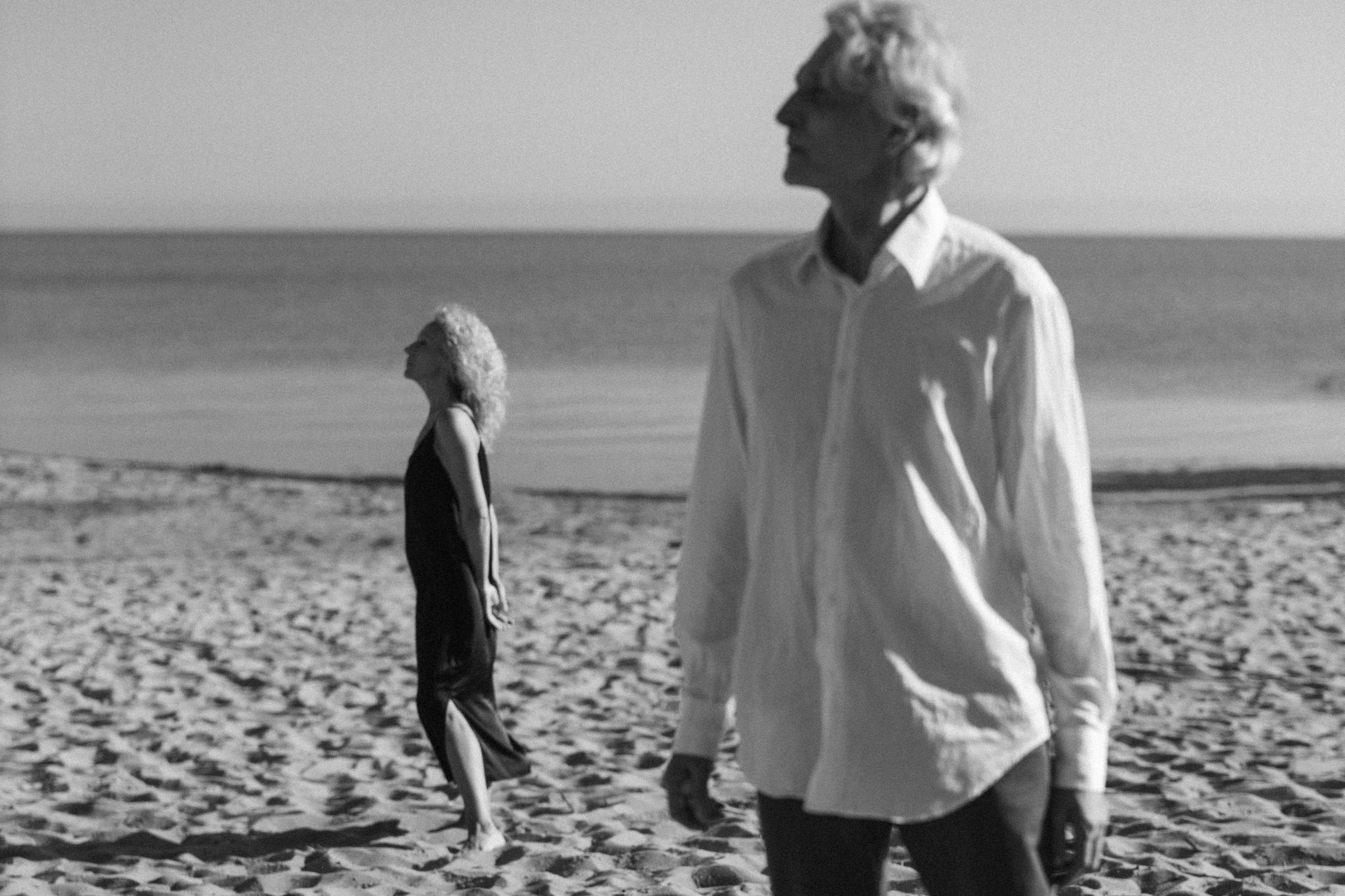 Older couple standing apart on a quiet beach, looking reflective under soft sunlight.