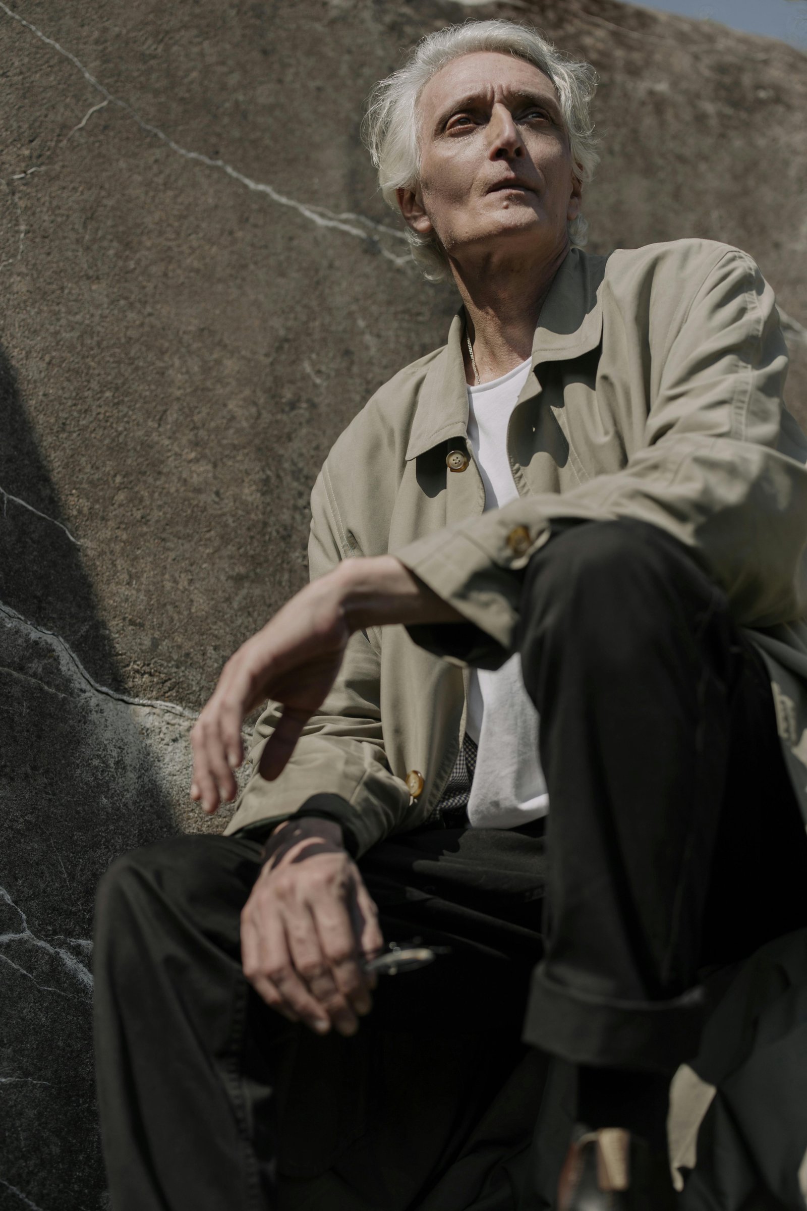 Older man sitting outdoors against a stone wall, looking reflective and thoughtful in natural light