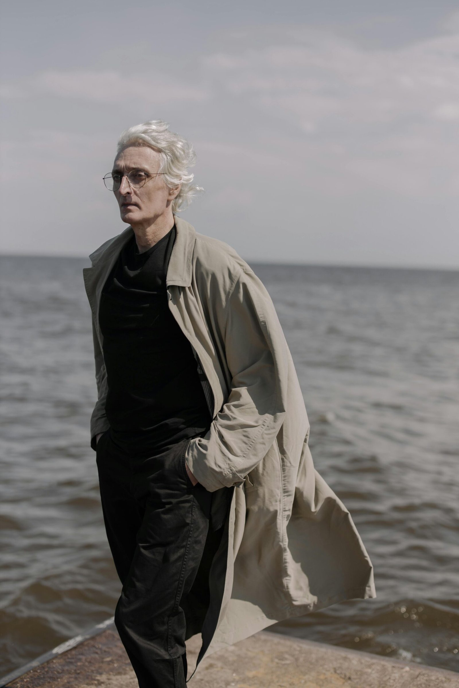 Older man walking alone by the ocean, wearing a long coat and glasses