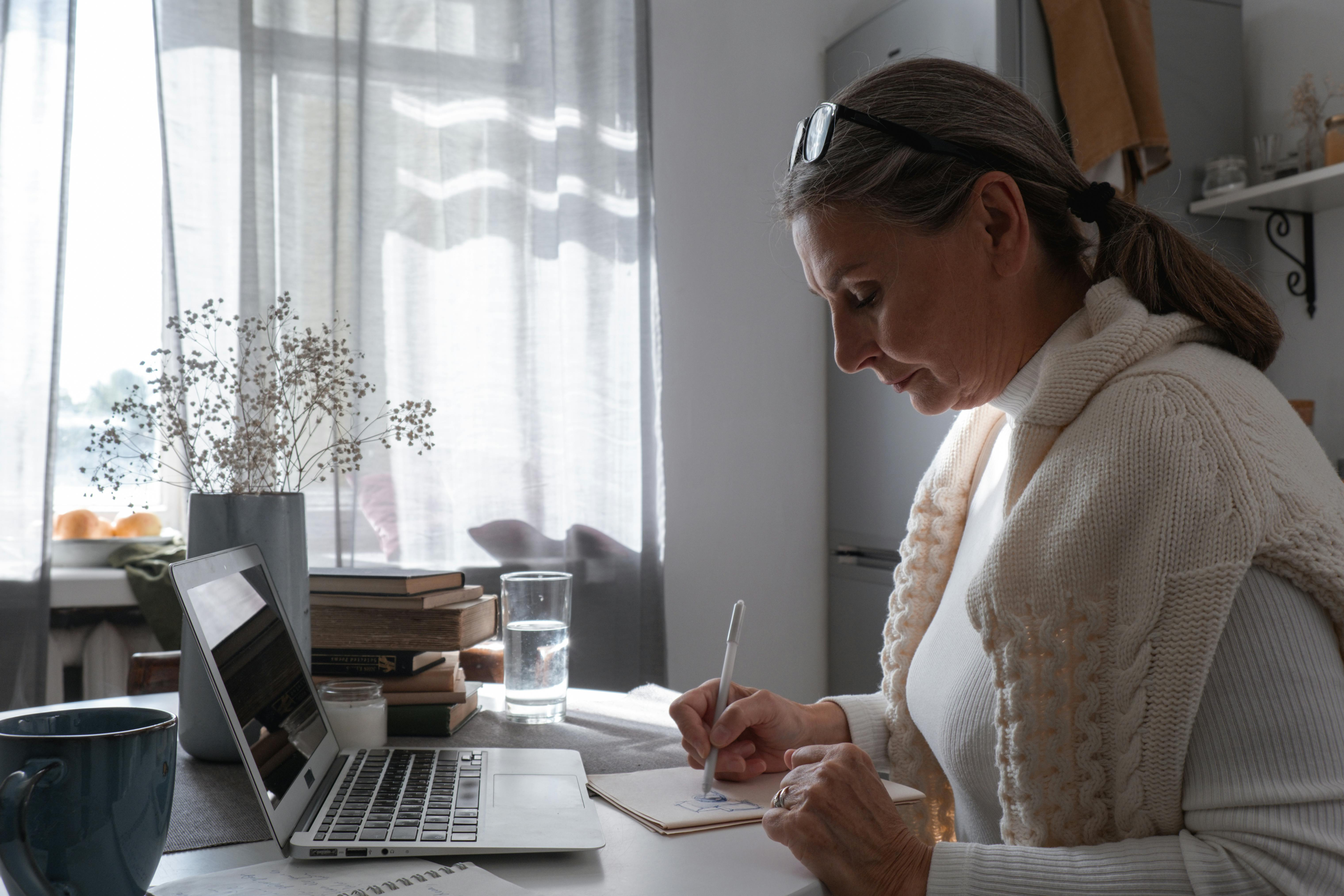 Woman over 40 working at a desk, writing notes beside a laptop in a calm home setting