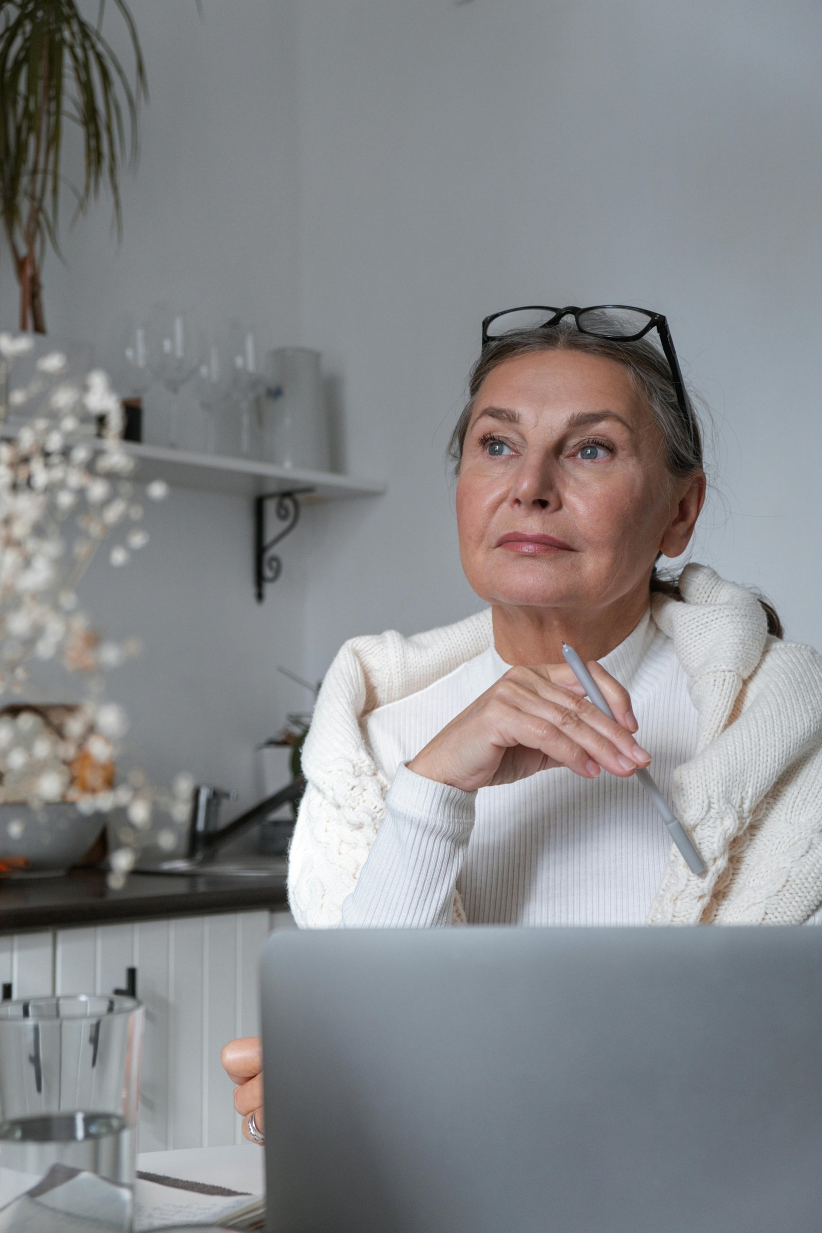 Thoughtful woman over 40 holding a pen while looking at a laptop in a bright kitchen workspace
