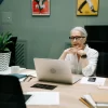 An older woman with gray hair and glasses works on a laptop at a modern office desk.