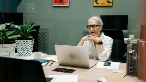 An older woman with gray hair and glasses works on a laptop at a modern office desk.