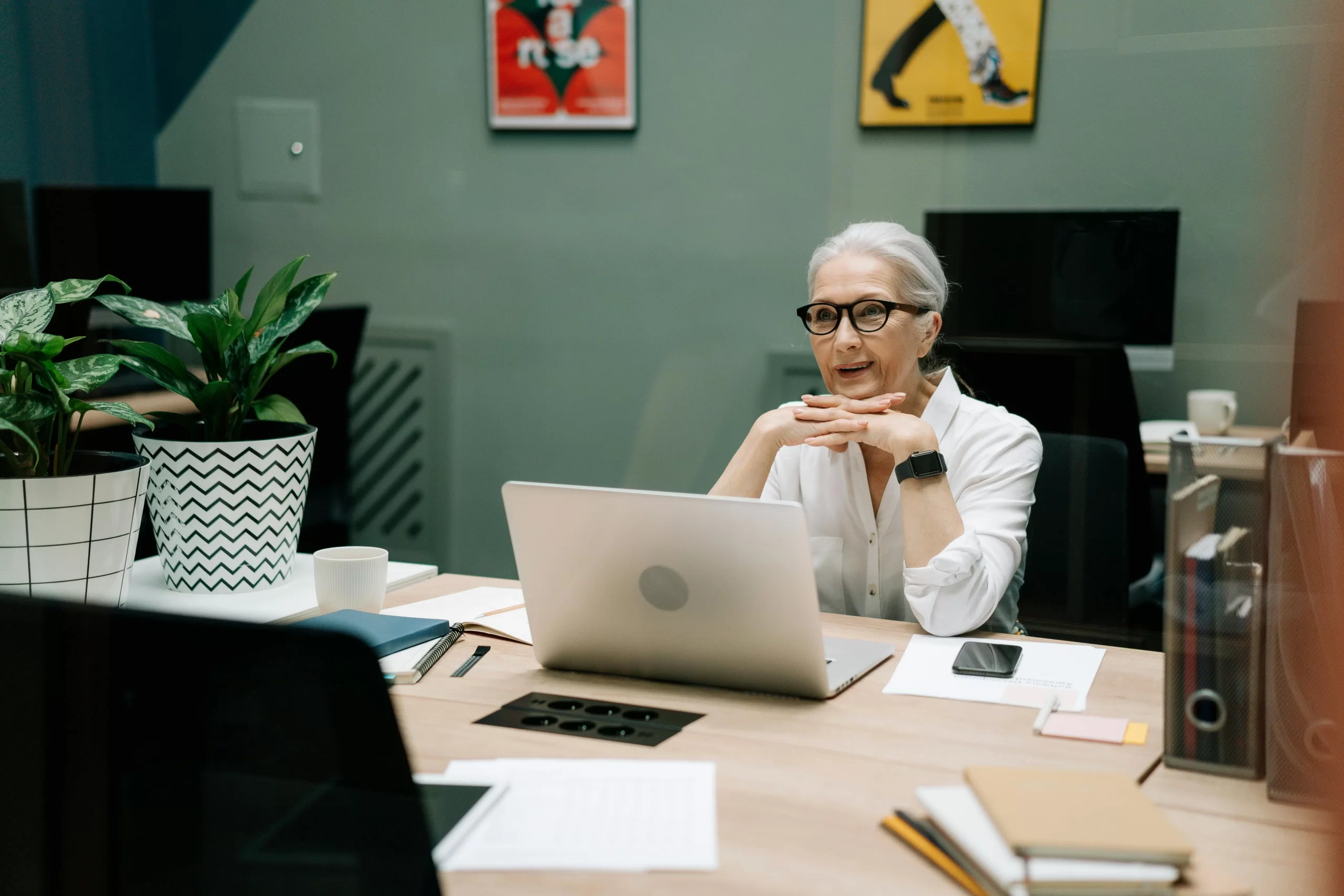 An older woman with gray hair and glasses works on a laptop at a modern office desk.