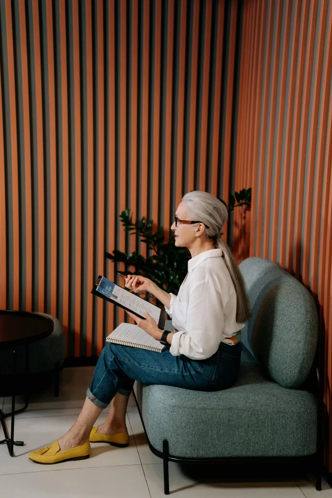 A senior woman with long gray hair reads from a clipboard while sitting on a modern sofa in a striped room.