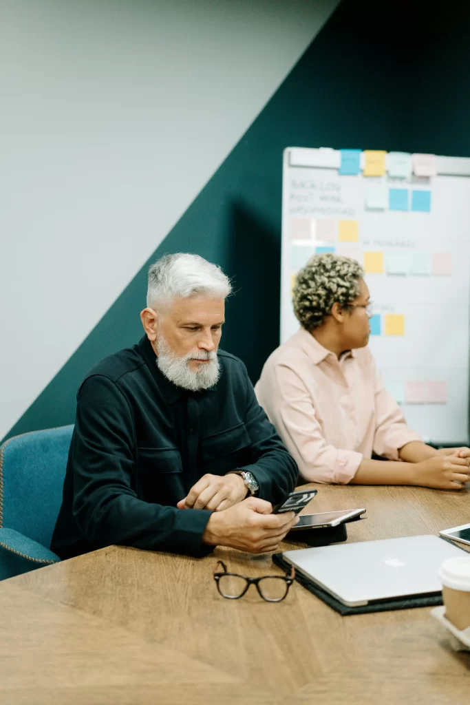 An older man with white hair checks his smartphone during a meeting beside coworkers in a conference room.