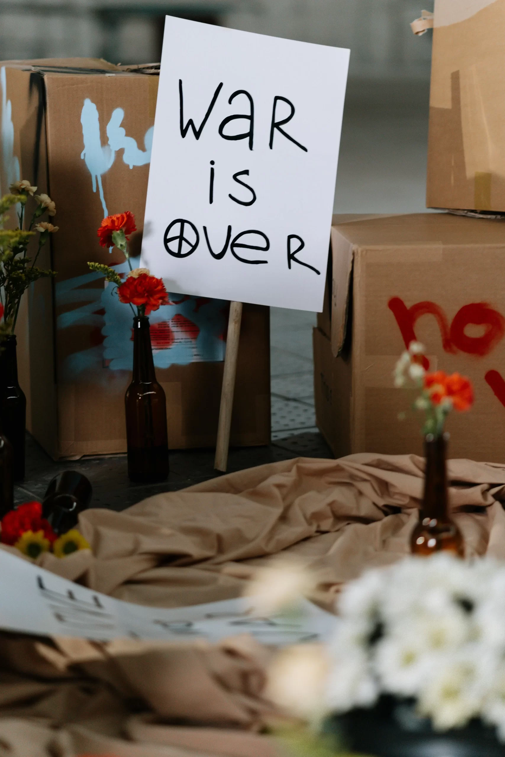 Peaceful protest setup with a handwritten ‘War Is Over’ sign surrounded by flowers and cardboard boxes.