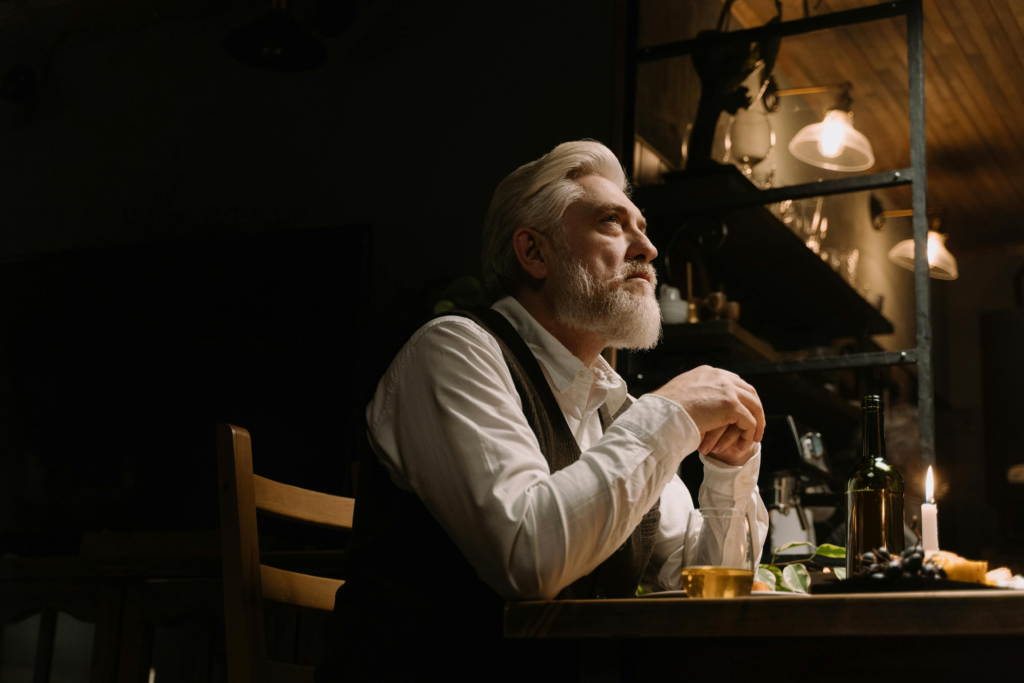Middle-aged man with gray hair sitting at a table with a drink, hands clasped, appearing deep in thought