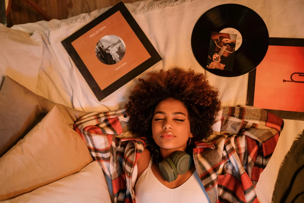 A young woman lying on a bed with vinyl records around her, wearing headphones around her neck.