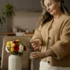 Older woman watering a small bouquet of flowers while wearing a neutral-toned cardigan, representing a calm, minimalist lifestyle.