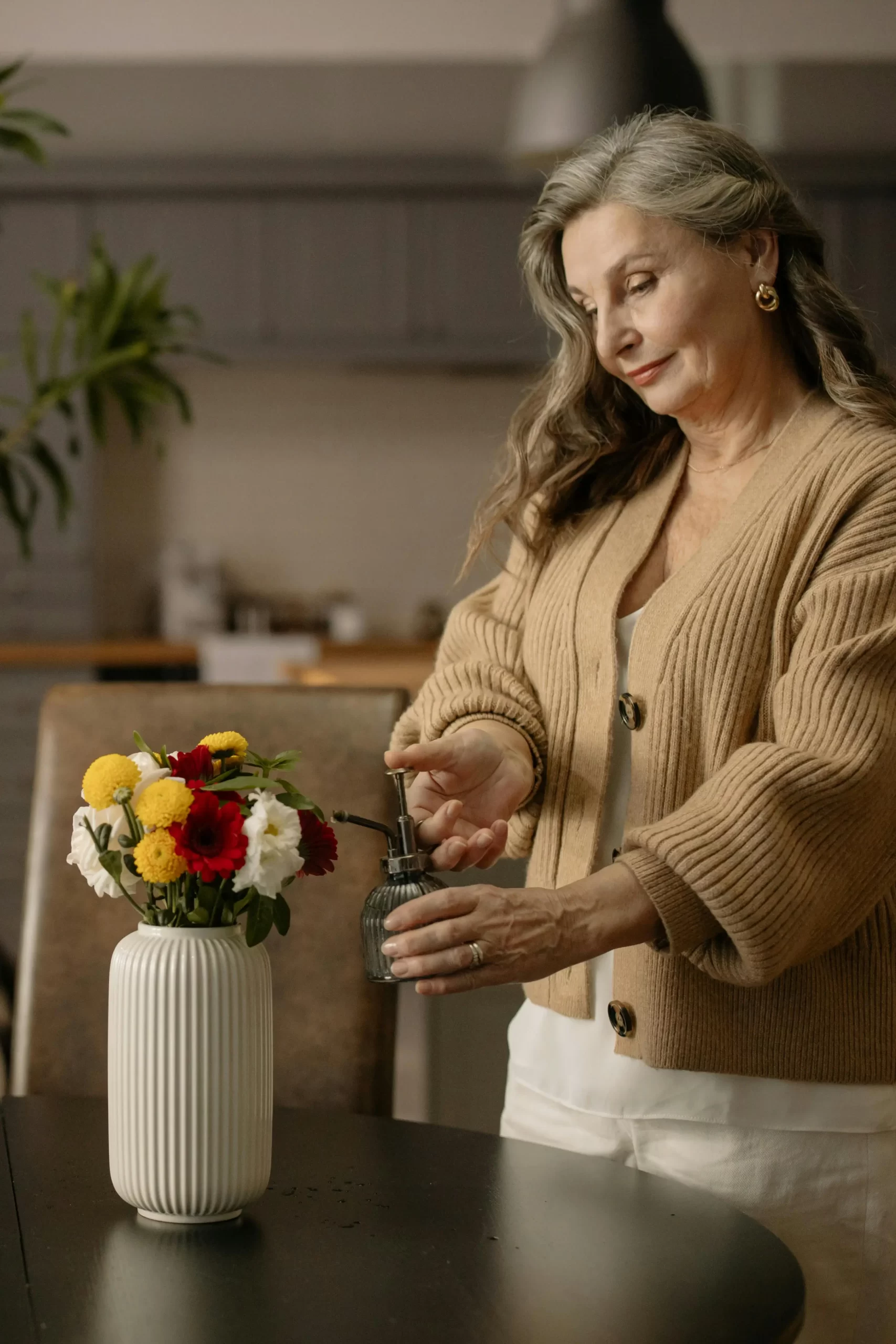Older woman watering a small bouquet of flowers while wearing a neutral-toned cardigan, representing a calm, minimalist lifestyle.