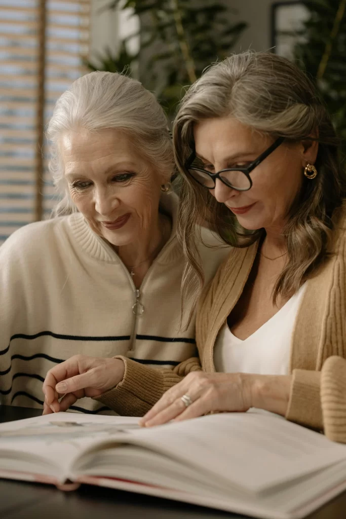 Two older women reading a book together at a table, both dressed in soft, classic wardrobe pieces.