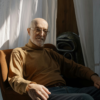 Older man sitting in a chair near a window, looking thoughtful during a quiet moment at home
