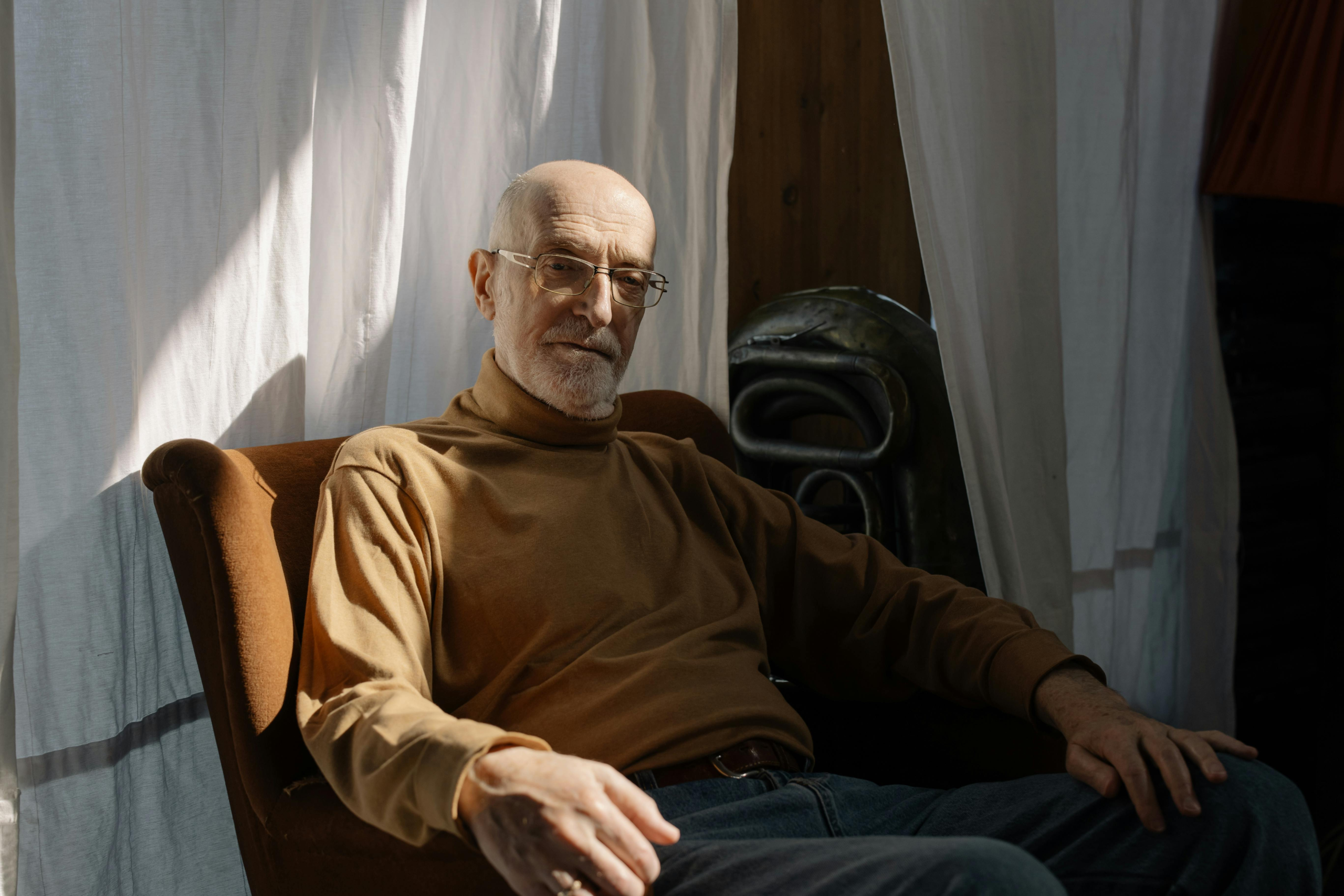 Older man sitting in a chair near a window, looking thoughtful during a quiet moment at home