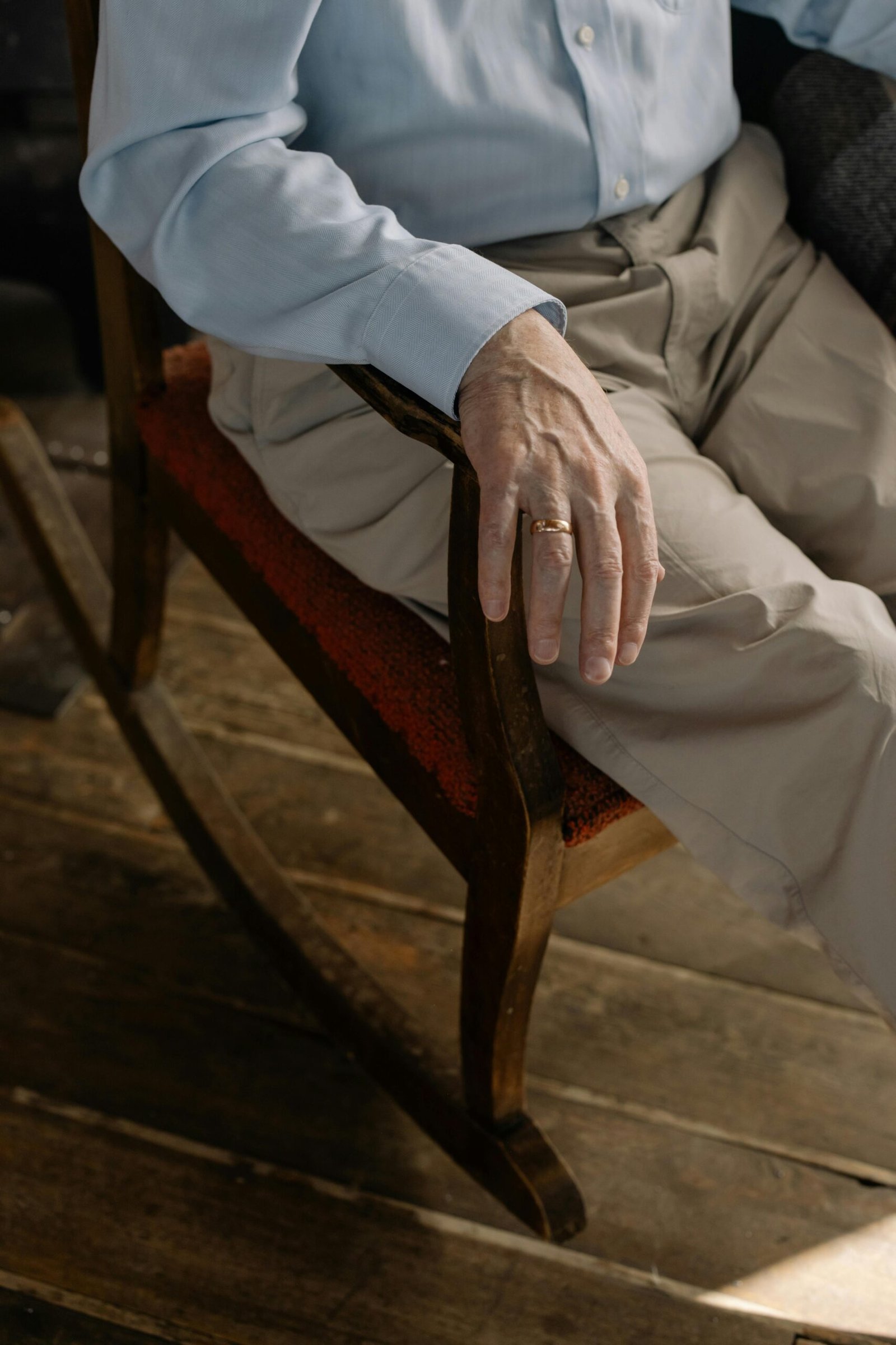 Close-up of an older man’s hand resting on a wooden rocking chair, showing visible aging and a gold wedding ring.