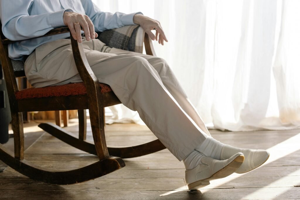 Senior man sitting in a wooden rocking chair with legs extended, relaxing in natural sunlight at home.