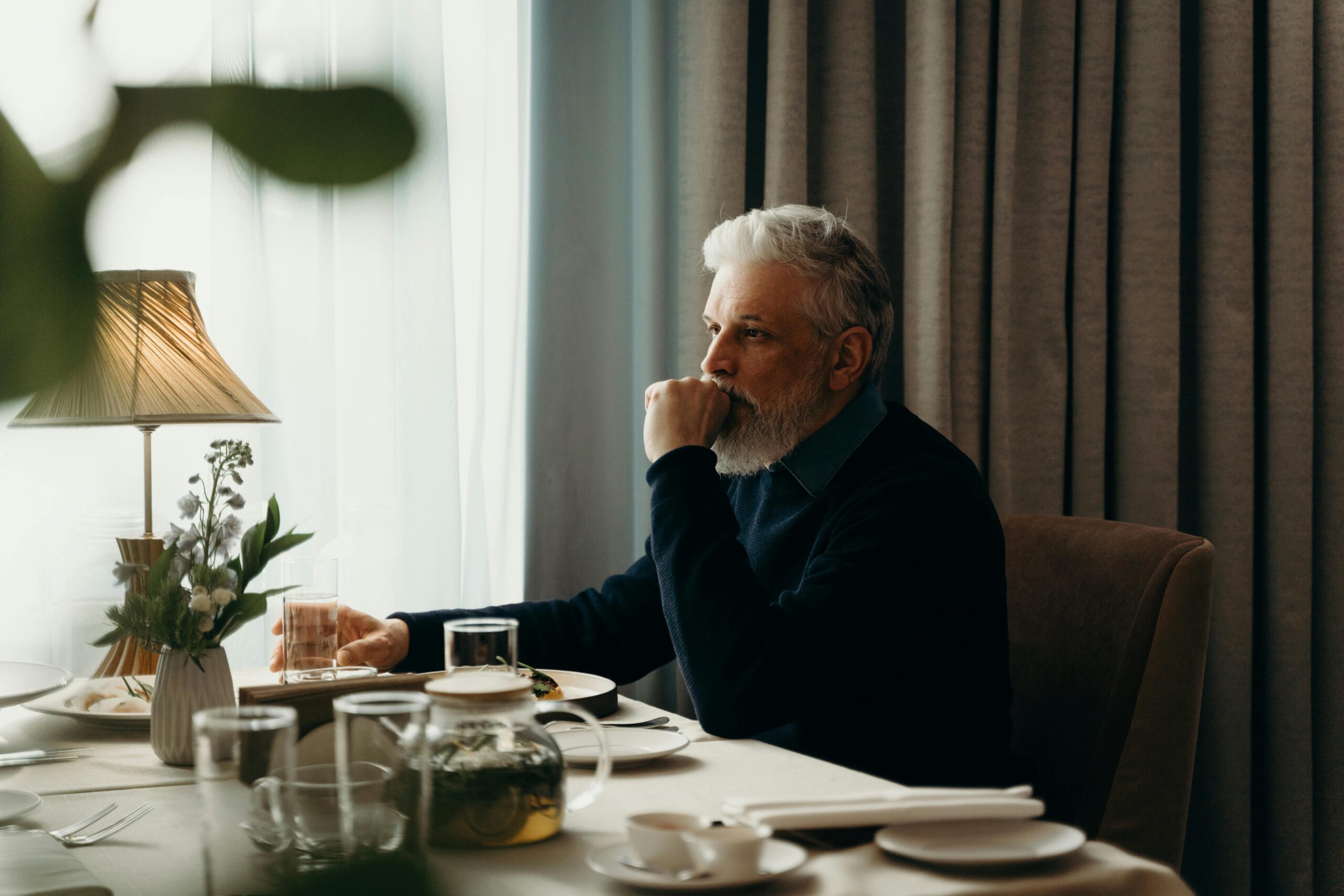 Man in his 60s sitting at a dining table, reflecting quietly in a calm, minimalist home
